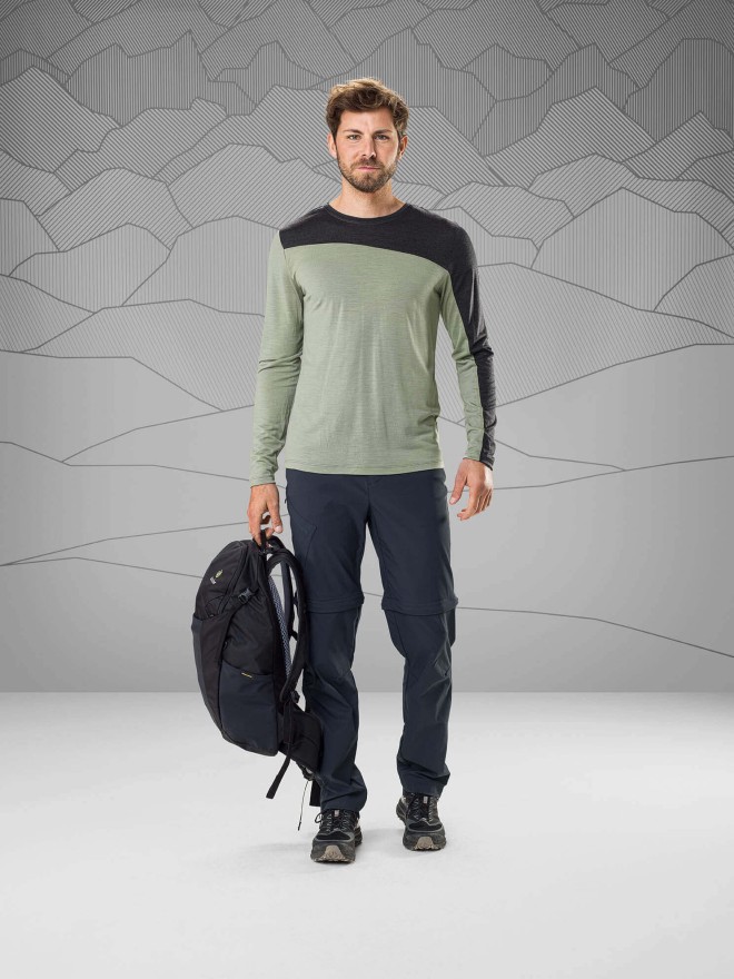 Men wearing a gray and green shirt with long sleeves and a dark gray trekking tousers in front of a gray background holding a black backpack in his right hand