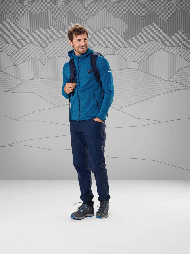 Man wearing a blue jacket and dark blue trekking pants standing in front of a gray background