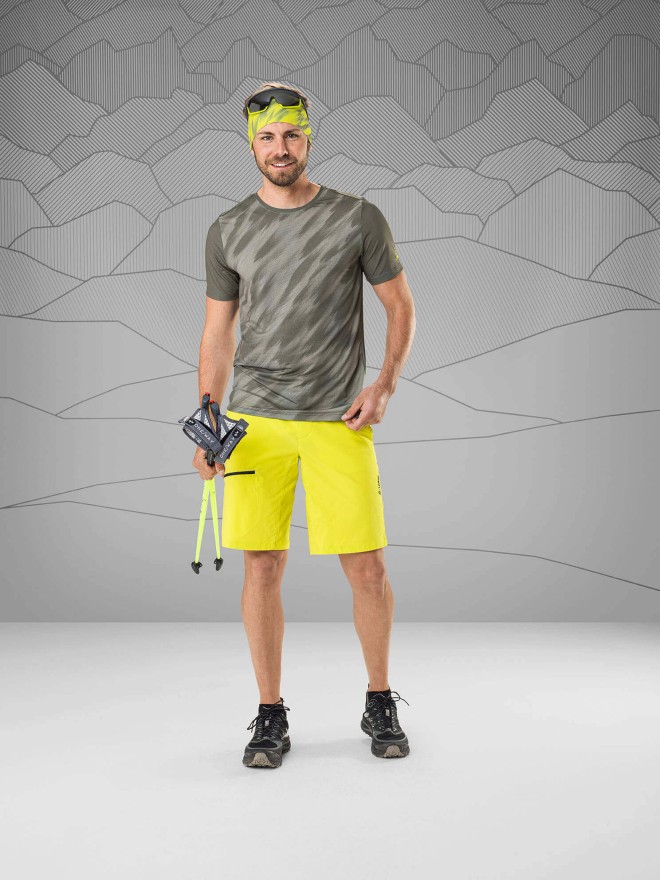 Man wearing a green/gray shirt and neon yellow shorts standing in front of a gray background